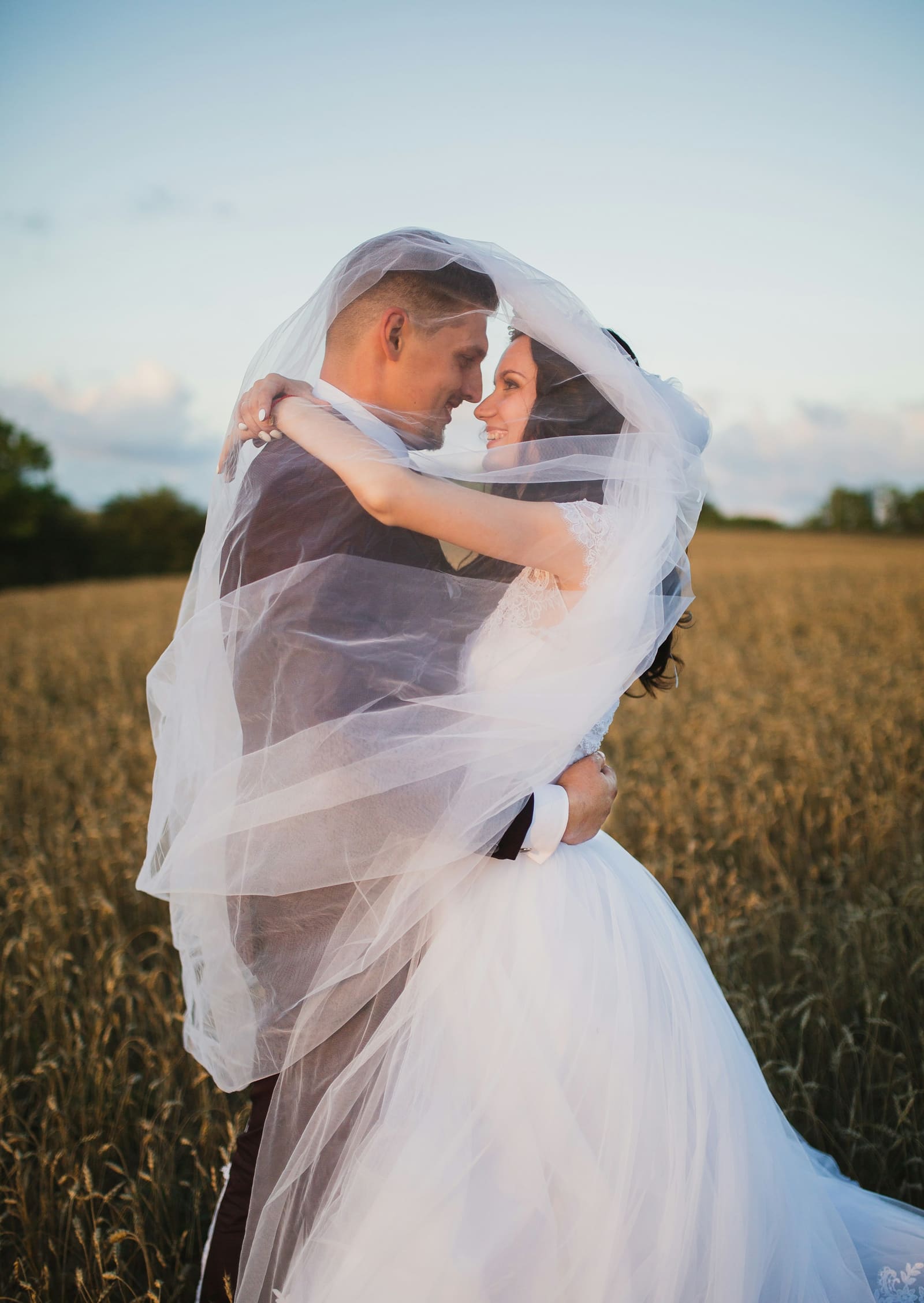 Bride and groom embracing under a flowing veil in a golden field at sunset
